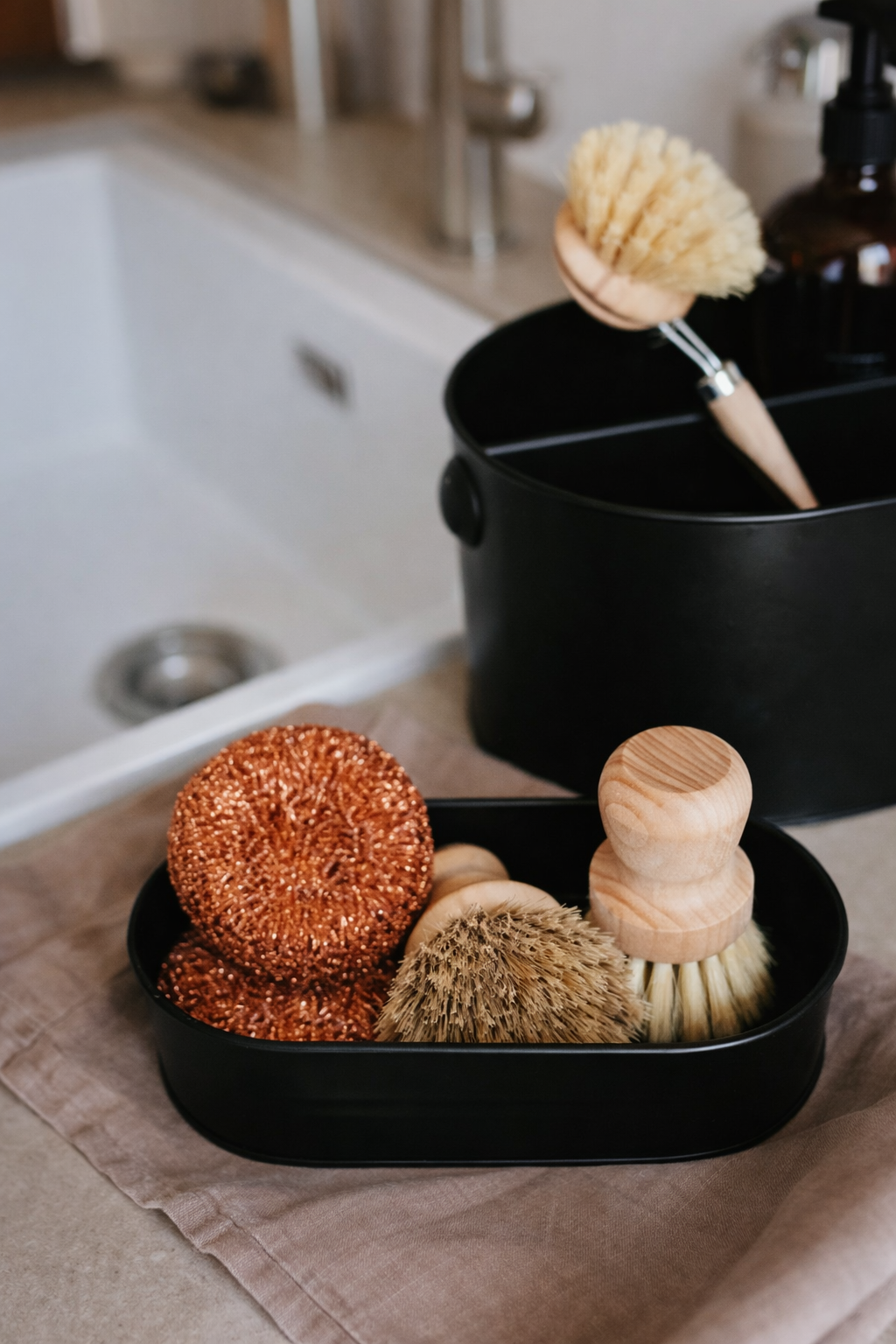 Natural sponges and wooden dish scrub brushes with copper scouring pads displayed near a kitchen sink, eco-friendly kitchen cleaning tools in a minimalist home interior with soft natural light.